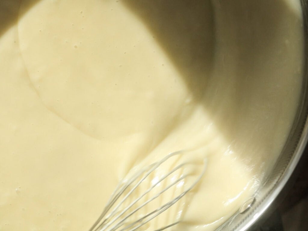 A metal whisk is partially submerged in a smooth, creamy bechamel inside a mixing bowl, with sunlight casting a shadow over the surface.